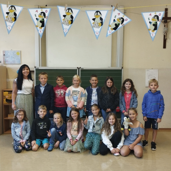 Eine Gruppe von Kindern und einer erwachsenen Frau posiert für ein Foto in einem Klassenzimmer. Ein Pinguinbanner hängt über ihnen.