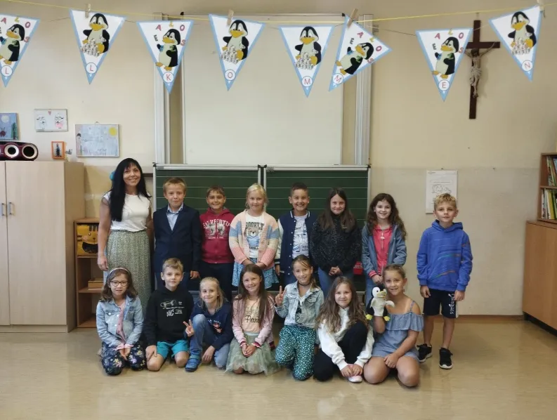 Eine Gruppe von Kindern und einer erwachsenen Frau posiert für ein Foto in einem Klassenzimmer. Ein Pinguinbanner hängt über ihnen.
