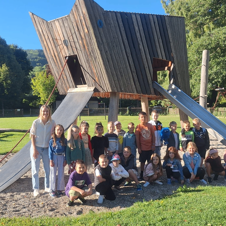 Eine Gruppe von Kindern und einer Frau posiert für ein Foto vor einem hölzernen Spielhaus mit Rutschen. Die Frau steht vorne, und die Kinder sitzen und knien auf dem Boden.
