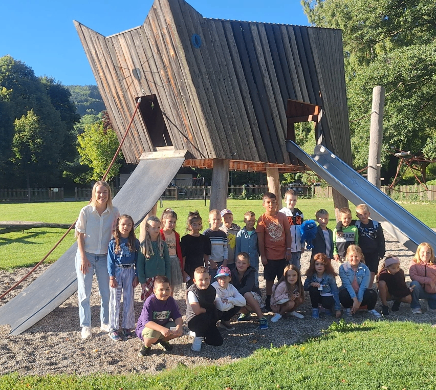 Eine Gruppe von Kindern und einer Frau posiert für ein Foto vor einem hölzernen Spielhaus mit Rutschen. Die Frau steht vorne, und die Kinder sitzen und knien auf dem Boden.