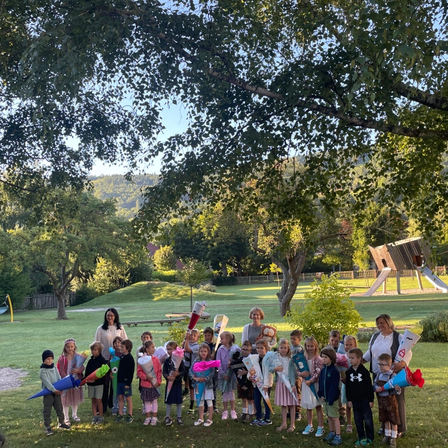 Eine Gruppe von Kindern und Erwachsenen steht in einem Park unter einem Baum mit einer Rutsche im Hintergrund. Sie halten Banner und Blumen.
