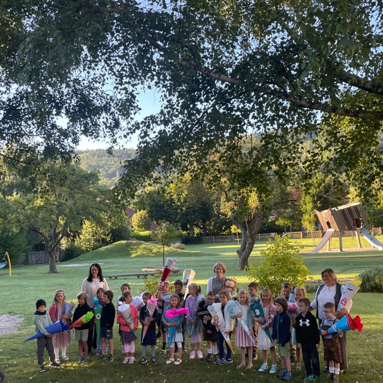 Eine Gruppe von Kindern und Erwachsenen steht in einem Park unter einem Baum mit einer Rutsche im Hintergrund. Sie halten Banner und Blumen.