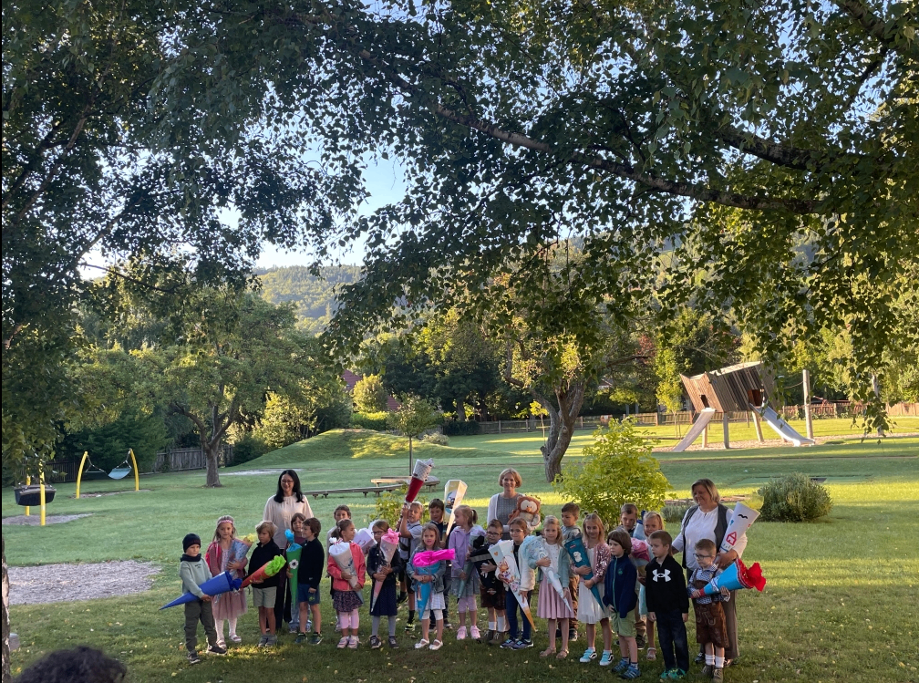 Eine Gruppe von Kindern und Erwachsenen steht in einem Park unter einem Baum mit einer Rutsche im Hintergrund. Sie halten Banner und Blumen.
