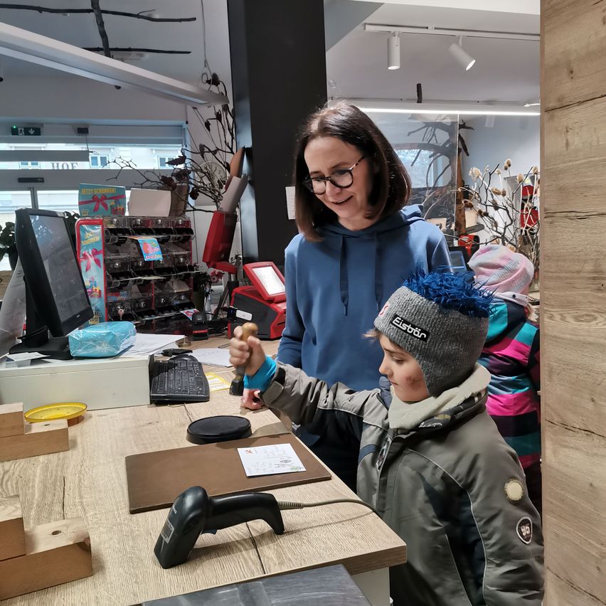 Eine Frau und ein kleiner Junge stehen lächelnd an einer Theke in einem Geschäft. Der Junge hält einen Holzstempel.