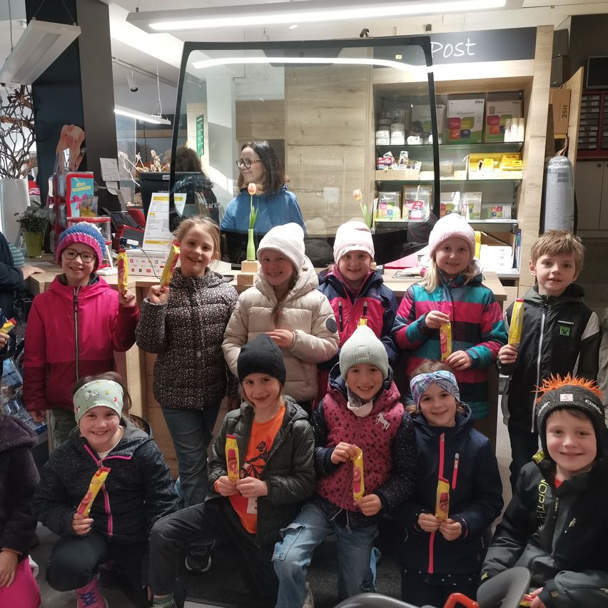 Eine Gruppe junger Kinder mit Mützen und Winterjacken, die lächelnd ein gelbes Objekt halten, posiert für ein Foto in einem Geschäft mit Theke und Regalen im Hintergrund.