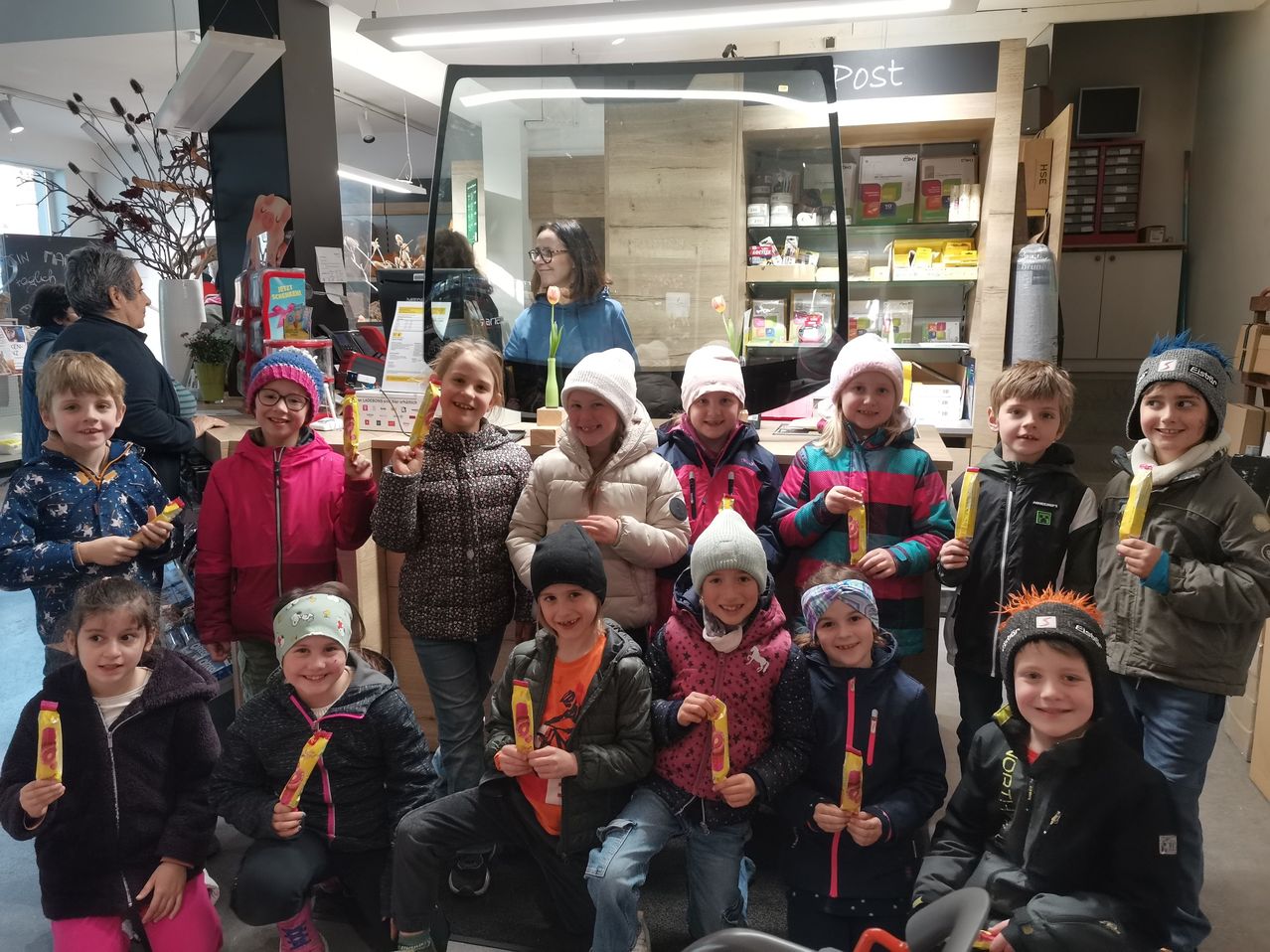 Eine Gruppe junger Kinder mit Mützen und Winterjacken, die lächelnd ein gelbes Objekt halten, posiert für ein Foto in einem Geschäft mit Theke und Regalen im Hintergrund.