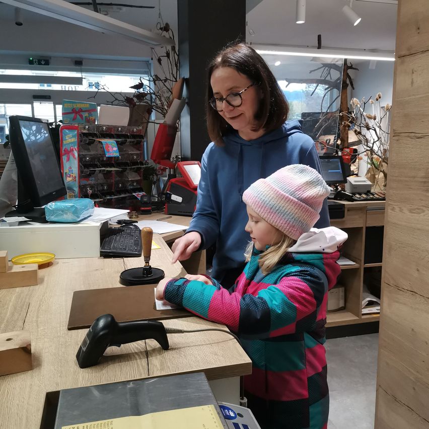 Eine Frau und ein Kind stehen vor einem Schreibtisch in einem Geschäft. Die Frau zeigt auf einen Stempel auf dem Schreibtisch.