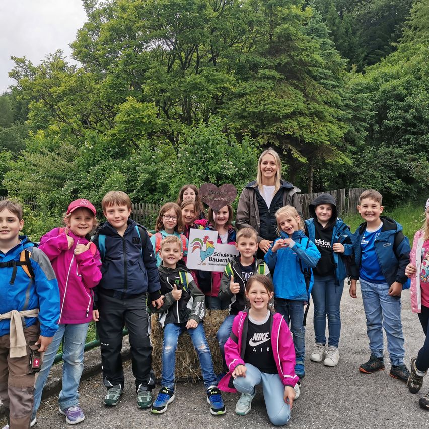 Eine Gruppe von Kindern und einer Frau posiert für ein Foto in einem bewaldeten Gebiet. Die Frau hält ein Schild mit einem Hahn und Text. Die Kinder tragen Rucksäcke und Jacken, und der Hintergrund ist üppig mit Grün bewachsen.