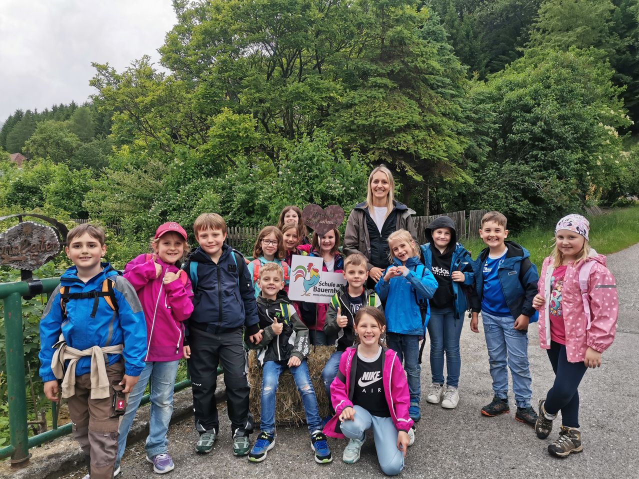 Eine Gruppe von Kindern und einer Frau posiert für ein Foto in einem bewaldeten Gebiet. Die Frau hält ein Schild mit einem Hahn und Text. Die Kinder tragen Rucksäcke und Jacken, und der Hintergrund ist üppig mit Grün bewachsen.