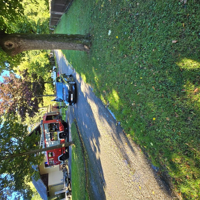 Luftaufnahme einer Einfahrt mit einem geparkten Auto und einem Feuerwehrfahrzeug. Der Bereich ist von saftigem, grünem Rasen und Bäumen umgeben, wobei ein Haus im Hintergrund zu sehen ist.