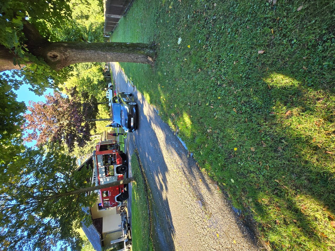 Luftaufnahme einer Einfahrt mit einem geparkten Auto und einem Feuerwehrfahrzeug. Der Bereich ist von saftigem, grünem Rasen und Bäumen umgeben, wobei ein Haus im Hintergrund zu sehen ist.