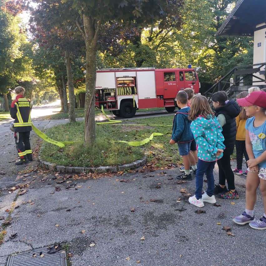 Ein Feuerwehrfahrzeug ist auf einer Straße geparkt, während Kinder beobachten. Ein Feuerwehrmann steht in der Nähe mit einem gelben Warnband um einen Baum.
