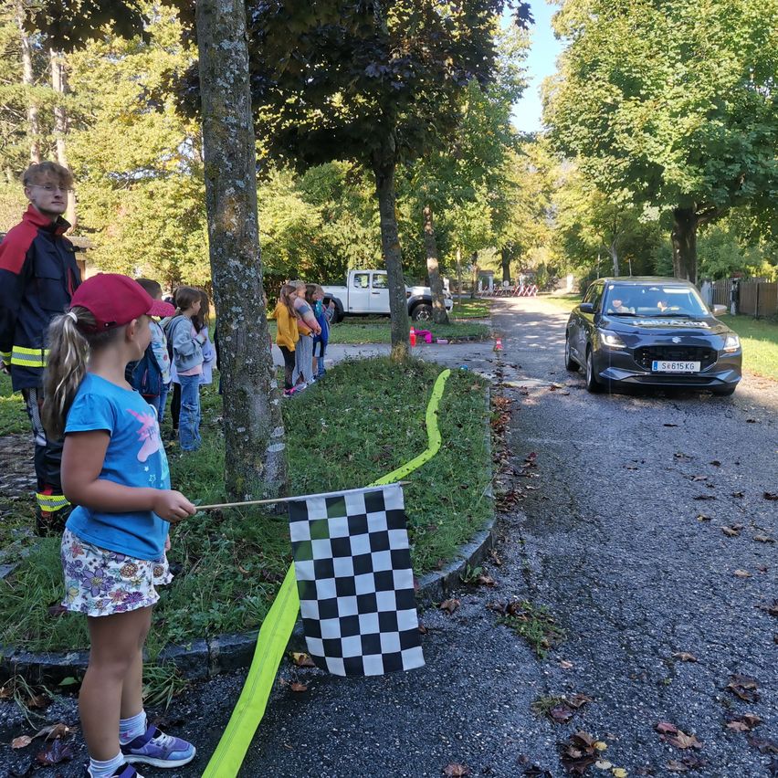Ein Mädchen hält eine gepunktete Flagge, während ein Auto die Straße hinunterfährt. Mehrere Kinder und Erwachsene beobachten von der Seite. Bäume säumen die Straße, und ein Pick-up-Truck ist in der Nähe geparkt.