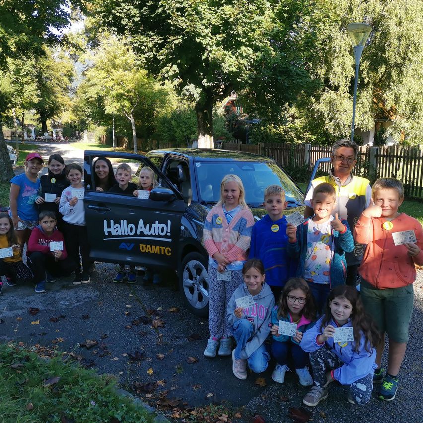 Eine Gruppe von Kindern und zwei Erwachsene posieren vor einem Auto mit dem Slogan 'Hallo Auto!'. Die Kinder halten Tickets und die Erwachsenen lächeln. Die Umgebung ist im Freien mit Bäumen und einem Weg.