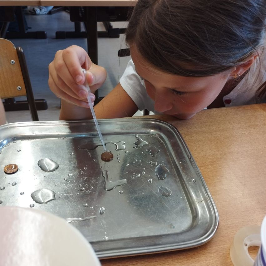 Ein junges Mädchen führt ein Experiment mit einer Pipette und Flüssigkeit auf einem Metalltablett durch, wobei Münzen und Wassertropfen sichtbar sind.