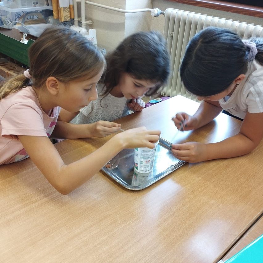 Drei Mädchen führen ein Experiment mit einer Dose, Münzen und einem Löffel auf einem Metalltablett an einem Schreibtisch durch.
