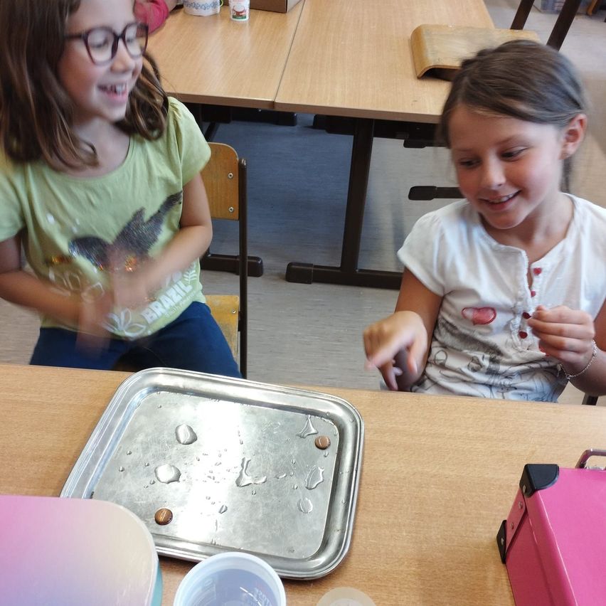 Zwei junge Mädchen sitzen an einem Tisch, lächeln und interagieren miteinander. Sie haben einen Metalltablett mit kleinen Gegenständen darauf, und es gibt verschiedene Dinge auf dem Tisch.