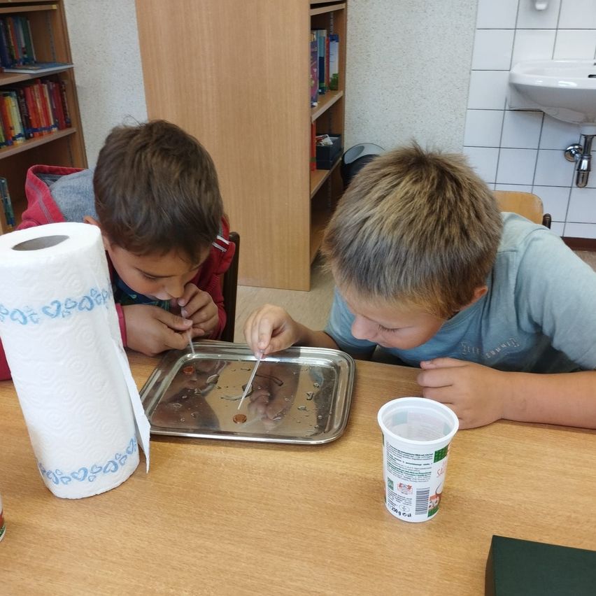 Zwei Jungen führen ein Experiment mit Münzen auf einem Tablett durch, ein Junge benutzt einen Strohhalm. Eine Papierhandtuchrolle und ein Becher stehen auf dem Tisch.