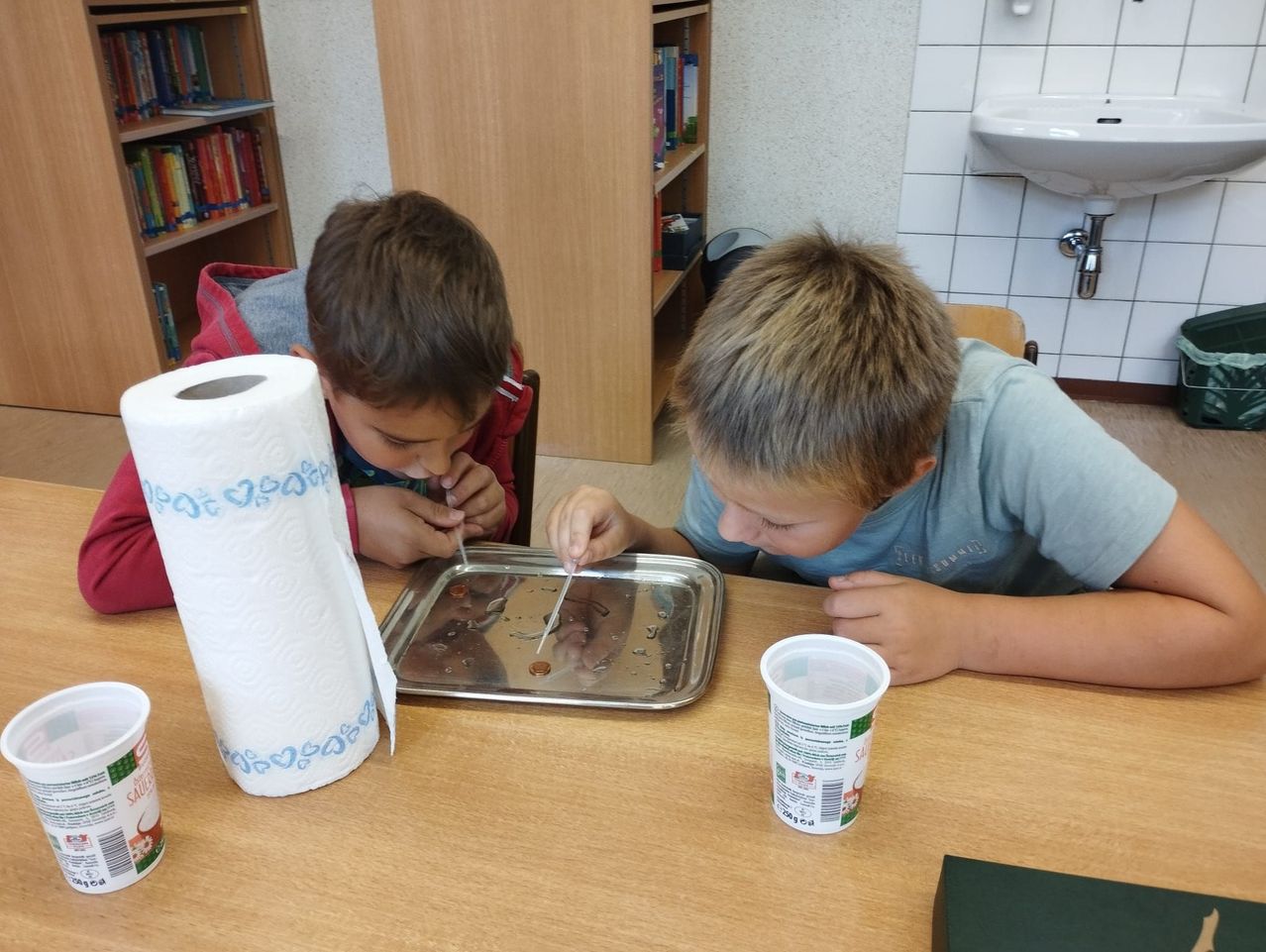 Zwei Jungen führen ein Experiment mit Münzen auf einem Tablett durch, ein Junge benutzt einen Strohhalm. Eine Papierhandtuchrolle und ein Becher stehen auf dem Tisch.