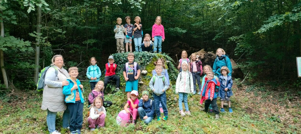 Eine Gruppe von Kindern, einige stehend und andere sitzend, posiert für ein Foto in einem Wald. Sie tragen verschiedene Jacken und Turnschuhe, einige haben Rucksäcke. Ein Baumstamm liegt im Hintergrund.