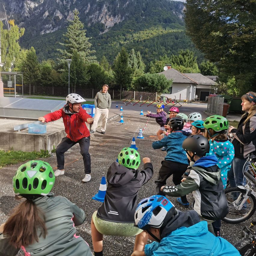 Kinder mit Helmen nehmen an einem Fahrradtraining auf einem Kiesplatz teil, mit Bergen im Hintergrund.