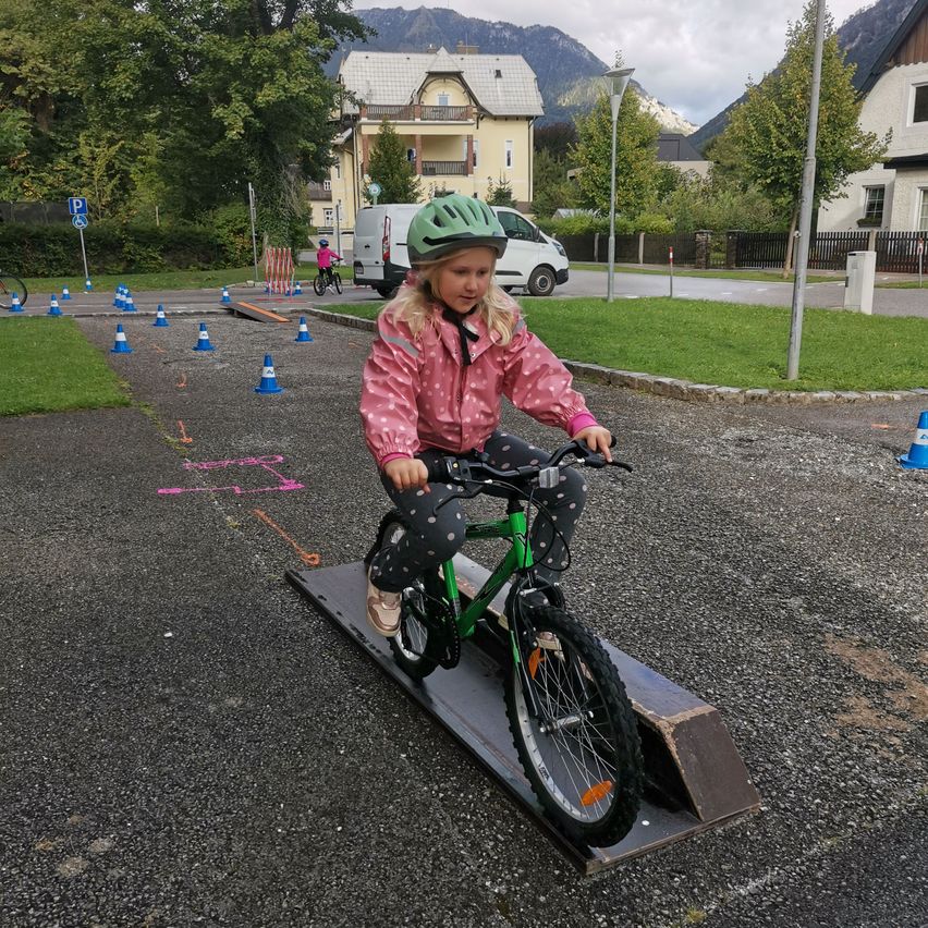 Ein junges Mädchen fährt ein grünes Fahrrad über eine Rampe in einem Park, umgeben von orangefarbenen Hütchen und Häusern.