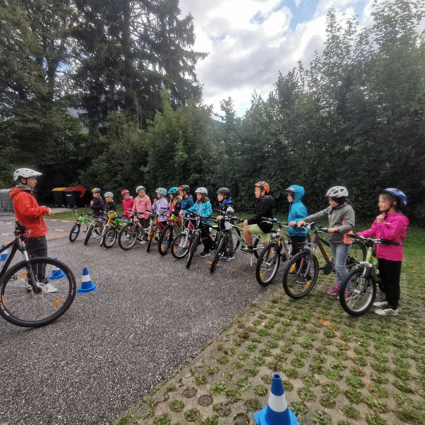 Eine Gruppe von Kindern, die Helme tragen, fährt auf einer Straße mit Fahrrädern. Sie stehen in einer Reihe und werden von einem Erwachsenen beaufsichtigt.