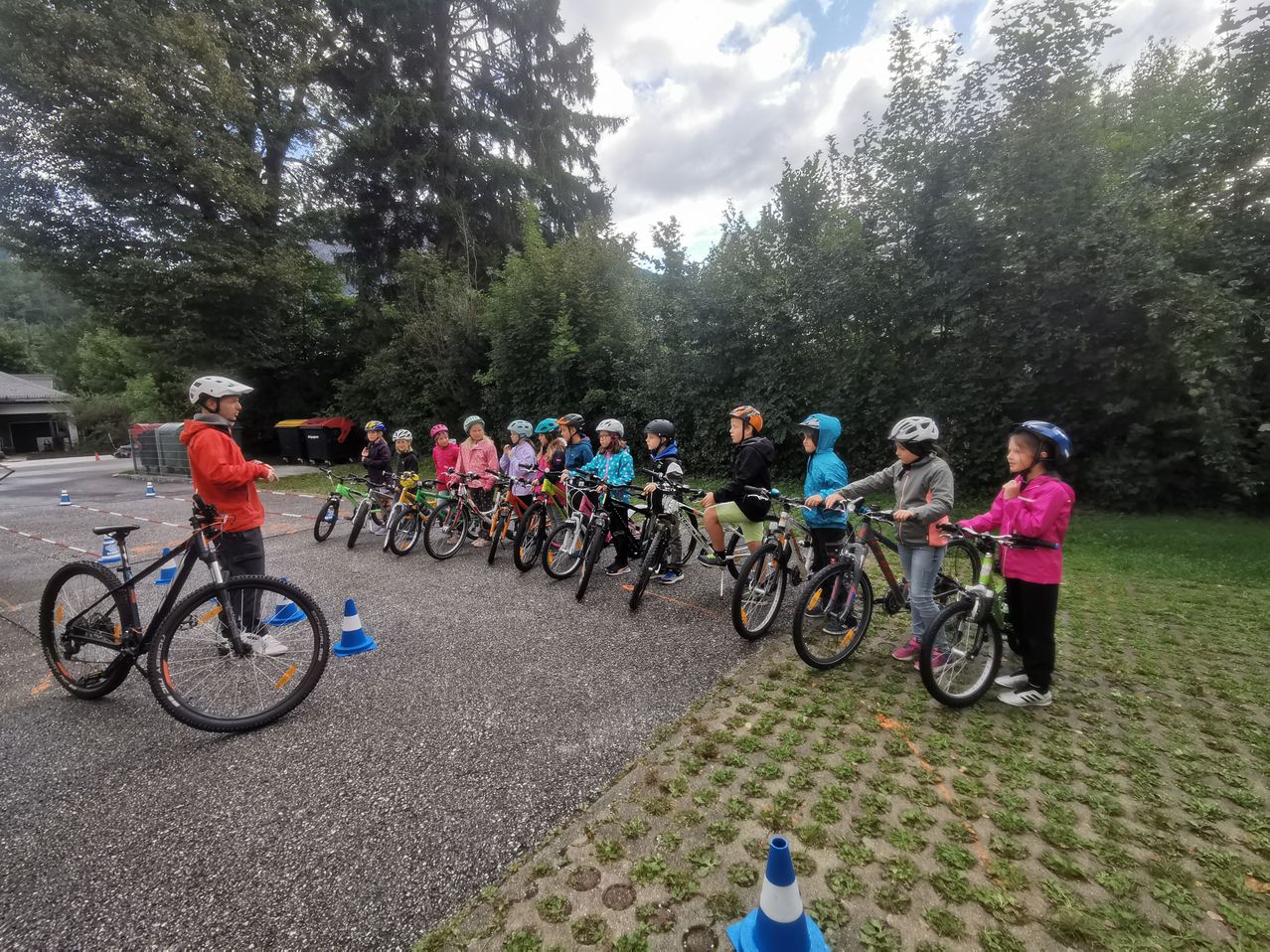 Eine Gruppe von Kindern, die Helme tragen, fährt auf einer Straße mit Fahrrädern. Sie stehen in einer Reihe und werden von einem Erwachsenen beaufsichtigt.