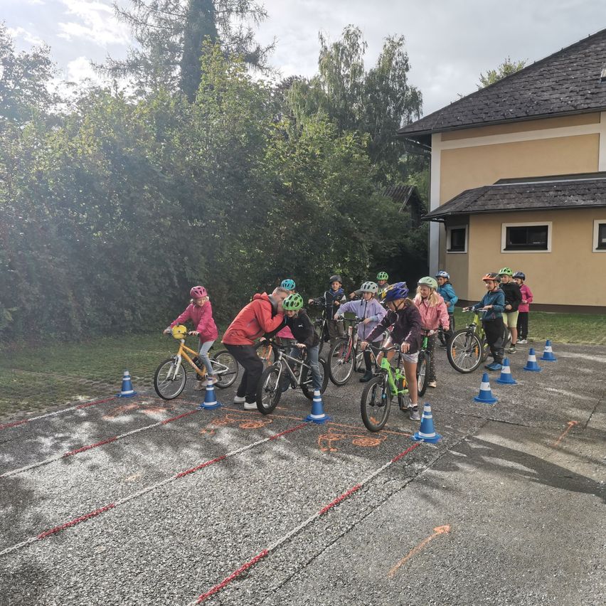 Eine Gruppe von Kindern in Helmen fährt auf einer asphaltierten Strecke mit Hütchen mit Fahrrädern. Sie sind im Freien in der Nähe eines Gebäudes mit schwarzem Dach.