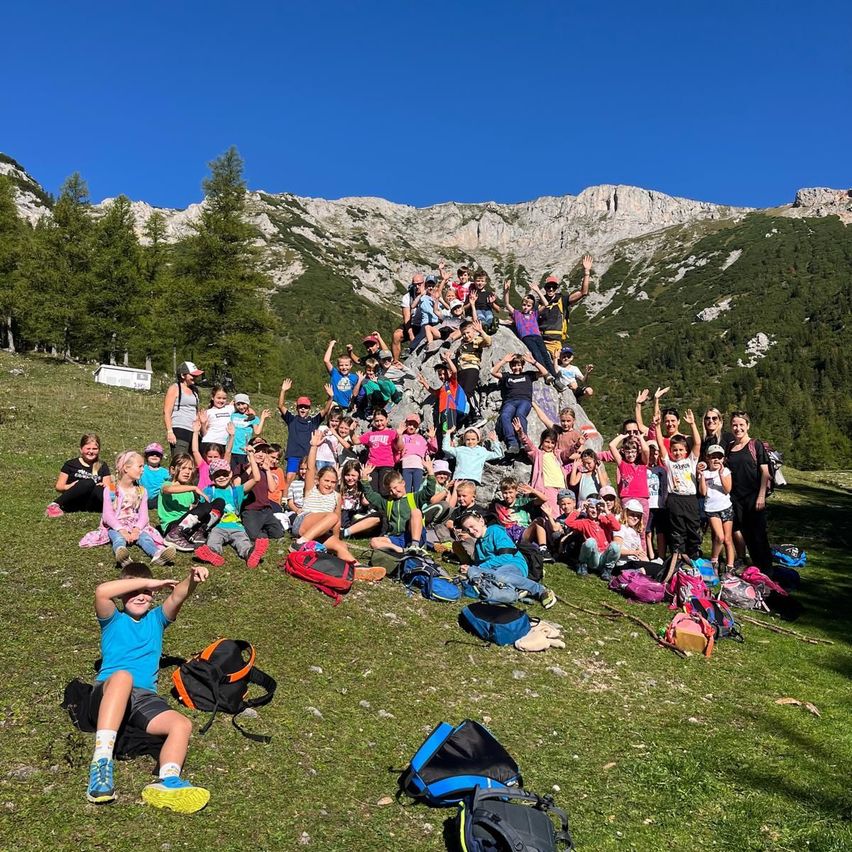 Eine Gruppe von Kindern und Erwachsenen in einem Berggebiet posiert für ein Foto. Einige sitzen, andere stehen auf einem Felsen. Sie haben verstreute Rucksäcke und Taschen.