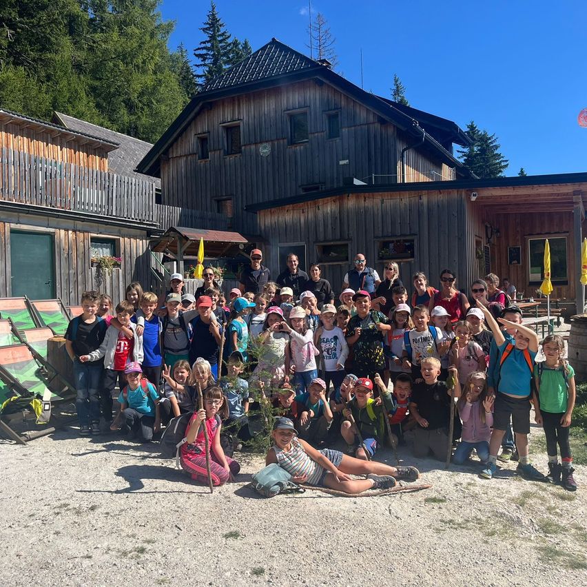 Eine Gruppe von Kindern und Erwachsenen posiert für ein Foto vor einem Holzgebäude mit vielen Fenstern und Bäumen im Hintergrund.