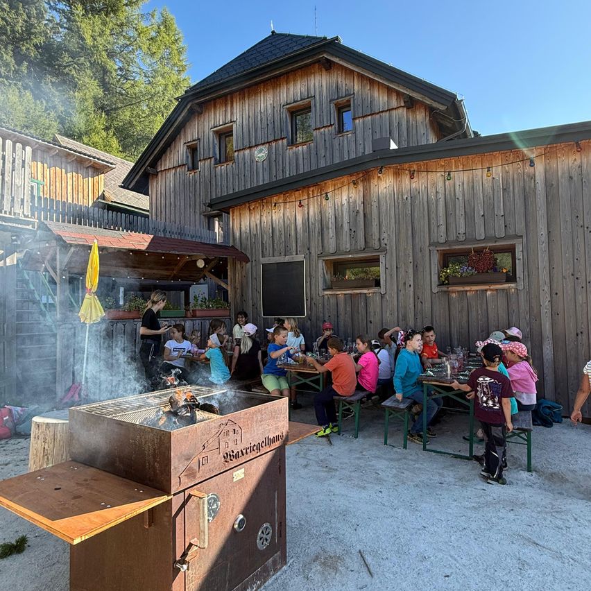 Eine Gruppe von Kindern und Erwachsenen sitzt um einen Tisch vor einem Holzgebäude herum, mit einem Grill in der Nähe, umgeben von Grün und einem klaren blauen Himmel.