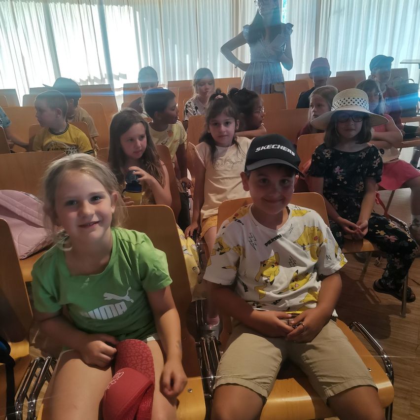 Eine Gruppe von Kindern sitzt in einem Klassenzimmer, mit einer Lehrerin im Hintergrund. Einige tragen Hüte, Brillen und Turnschuhe. Ein Junge lächelt und hält ein Handy.