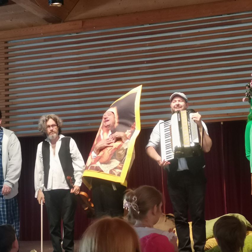 Eine Gruppe von Darstellern auf der Bühne, einer hält ein großes Plakat, ein anderer spielt Akkordeon, und ein junges Mädchen davor, während das Publikum zuschaut.