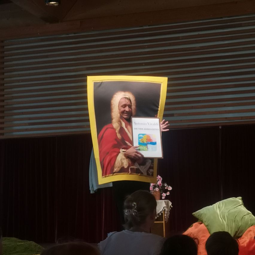 Eine Person hält ein gerahmtes Porträt von Antonio Vivaldi in einem Raum mit einer gestreiften Wand. Das Porträt zeigt Vivaldi in einem roten Gewand, das ein Buch hält. Vor dem Porträt befindet sich ein gelber Rahmen. Unten vor dem Porträt sind Blumen und Kissen zu sehen.