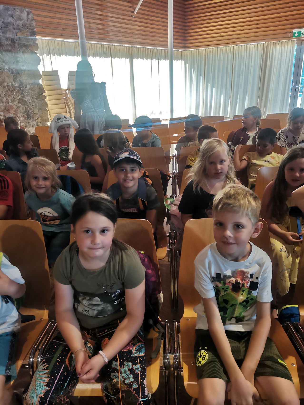 Eine Gruppe von Kindern sitzt in Stühlen in einem Raum. Sie tragen verschiedene Kleidungsstücke und einige haben Rucksäcke. Ein Mädchen in der vorderen Reihe lächelt, während andere in verschiedene Richtungen schauen.