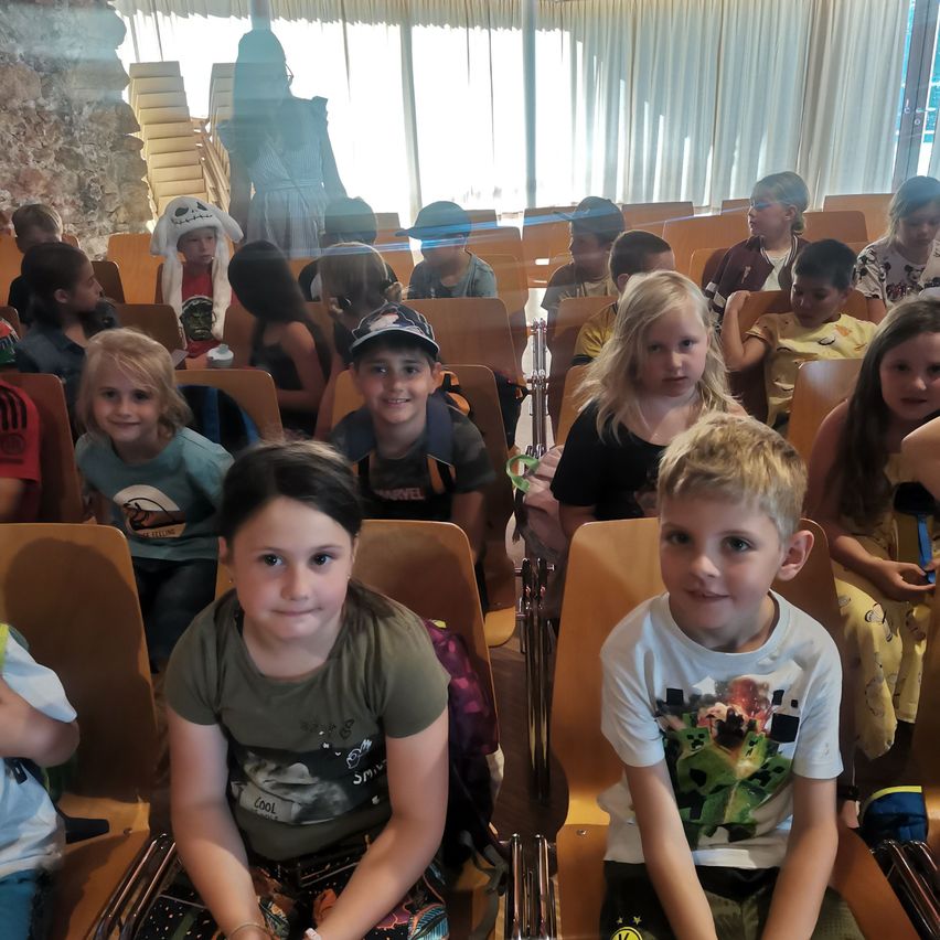 Eine Gruppe von Kindern sitzt in Stühlen in einem Raum. Sie tragen verschiedene Kleidungsstücke und einige haben Rucksäcke. Ein Mädchen in der vorderen Reihe lächelt, während andere in verschiedene Richtungen schauen.