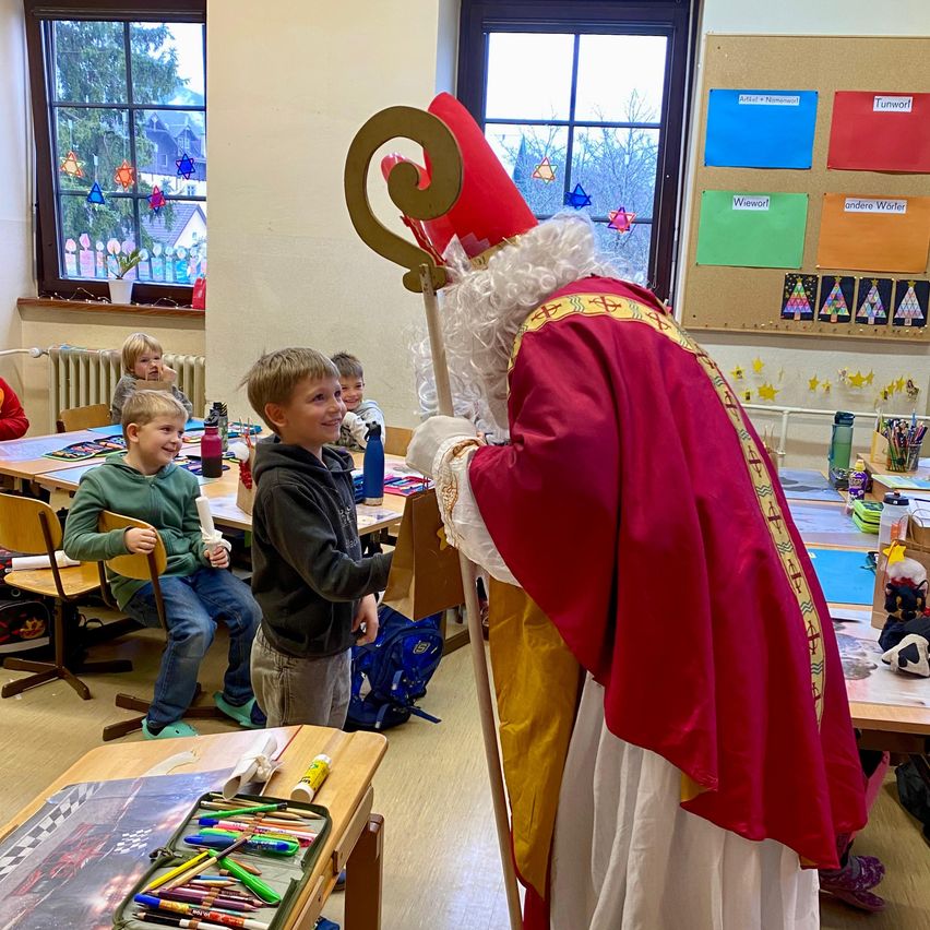Eine Person in Bischofskleidung mit einem goldenen Stab und einem roten Hut steht in einem Klassenzimmer mit Kindern, einer hält eine Flasche. Bunte Papiere sind an der Wand angeheftet.