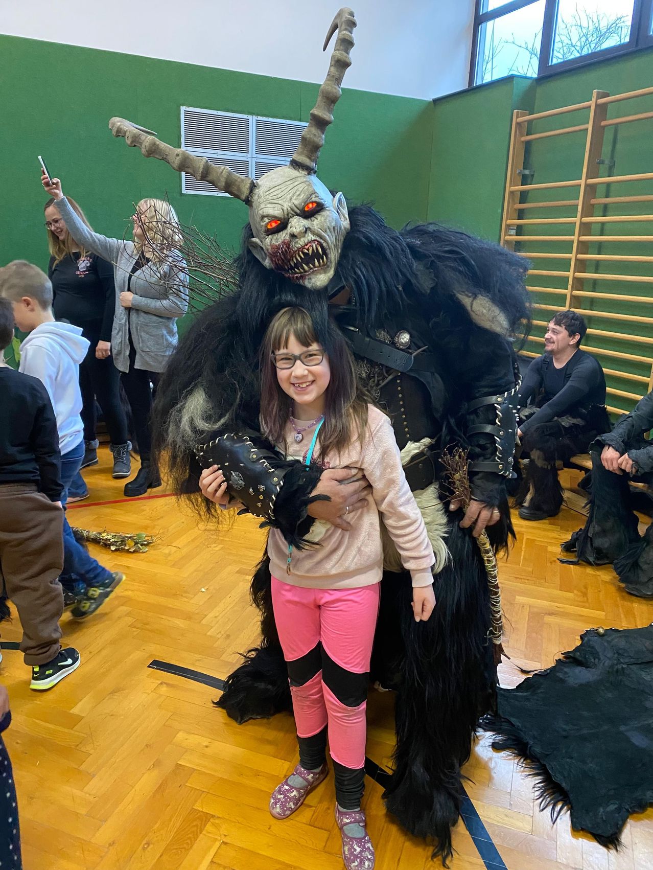 Ein Mädchen in rosa Hosen und einer Brille steht neben einer Person in einem gehörnten Monsterkostüm mit roten Augen und hält einen schwarzen Handschuh in einer Indoor-Umgebung mit Leuten um sie herum.