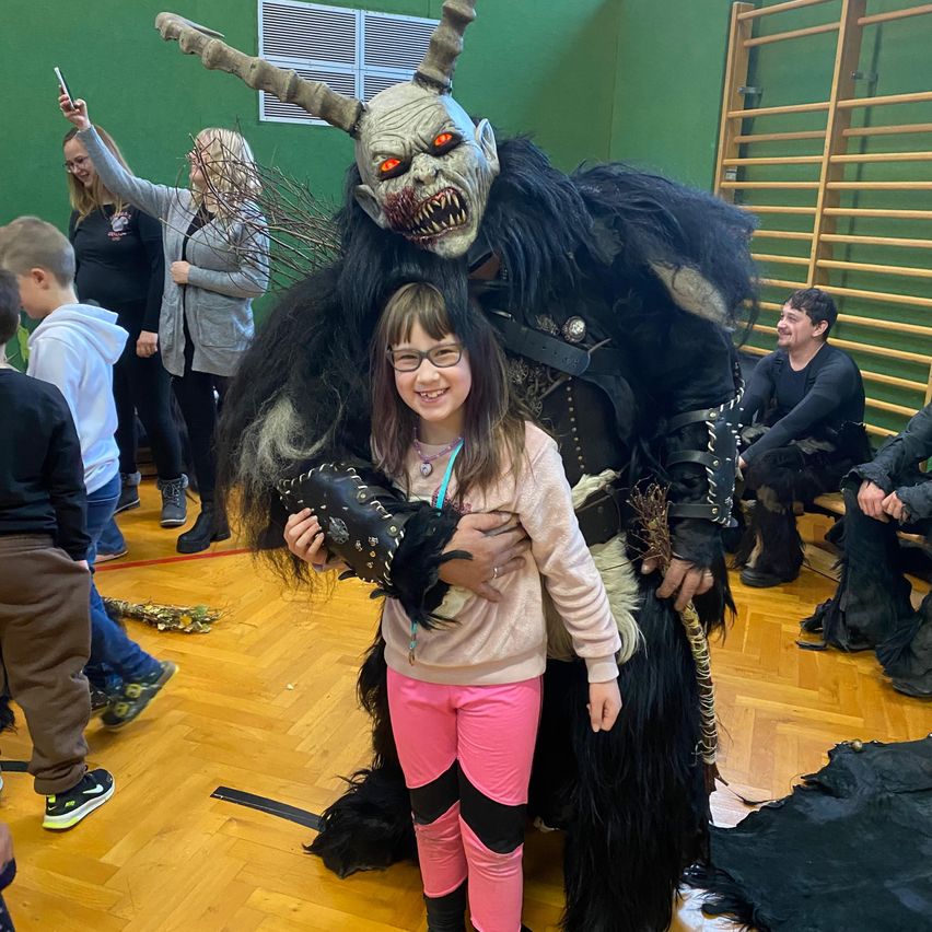 Ein Mädchen in rosa Hosen und einer Brille steht neben einer Person in einem gehörnten Monsterkostüm mit roten Augen und hält einen schwarzen Handschuh in einer Indoor-Umgebung mit Leuten um sie herum.