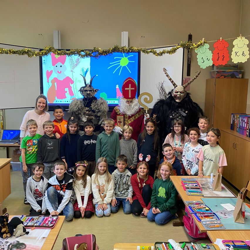 Eine Gruppe von Kindern in einem Klassenzimmer posiert für ein Foto. Sie tragen verschiedene Kostüme, darunter ein Weihnachtsmann-Kostüm. Dahinter zeigt eine Projektionsleinwand ein Cartoonbild. Auf der linken Seite steht eine Frau lächelnd neben einem Schreibtisch mit einem Laptop.