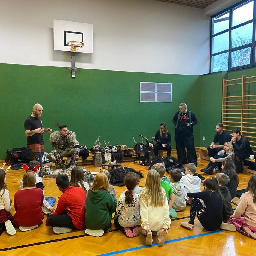 Ein Indoor-Basketballplatz mit zwei Männern, von denen einer Rentierhörner trägt, und Kindern, die vor ihnen sitzen. Die Wände sind grün und es gibt Fenster auf der rechten Seite.