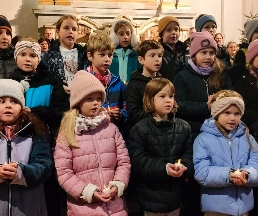 Eine Gruppe von Kindern in Winterkleidung, die Kerzen halten, stehen in einer Kirche zusammen. Sie scheinen an einer Zeremonie teilzunehmen.