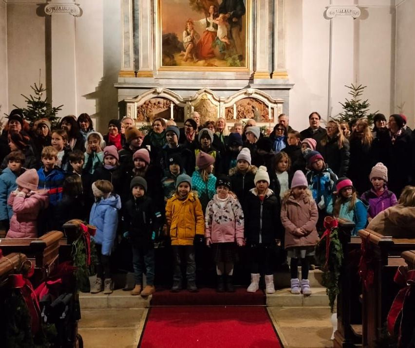 Eine Gruppe von Kindern und Erwachsenen versammelt sich in einer Kirche mit einem roten Teppich vor einem Altar und einem Gemälde von Jesus. Einige tragen Mützen und Stiefel.