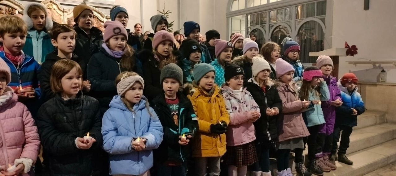 Eine Gruppe von Kindern mit brennenden Kerzen steht in einer Reihe, trägt Winterkleidung und Mützen. Hinter ihnen stehen Erwachsene, ebenfalls in warme Kleidung gehüllt. Die Umgebung scheint sich im Innenbereich zu befinden, mit einem Weihnachtsbaum und dekorativen Elementen.