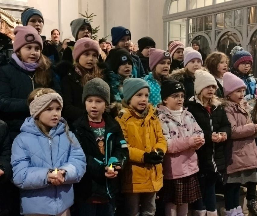 Eine Gruppe von Kindern mit brennenden Kerzen steht in einer Reihe, trägt Winterkleidung und Mützen. Hinter ihnen stehen Erwachsene, ebenfalls in warme Kleidung gehüllt. Die Umgebung scheint sich im Innenbereich zu befinden, mit einem Weihnachtsbaum und dekorativen Elementen.