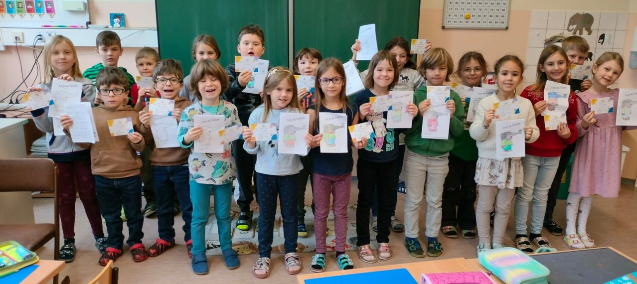 Eine Gruppe von Kindern steht in einem Klassenzimmer, hält Urkunden in den Händen und lächelt für ein Foto. Sie tragen Freizeitkleidung und Turnschuhe. An der Wand hinter ihnen ist eine grüne Tafel mit Zahlen angebracht.