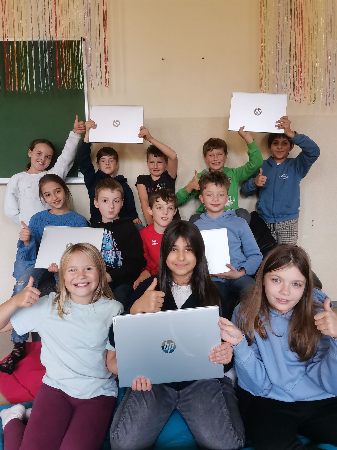 Eine Gruppe von Kindern im Klassenzimmer, die HP-Laptops halten, lächeln und Daumen hoch geben. Die Kinder sind in Freizeitkleidung gekleidet, einige sitzen auf dem Boden.