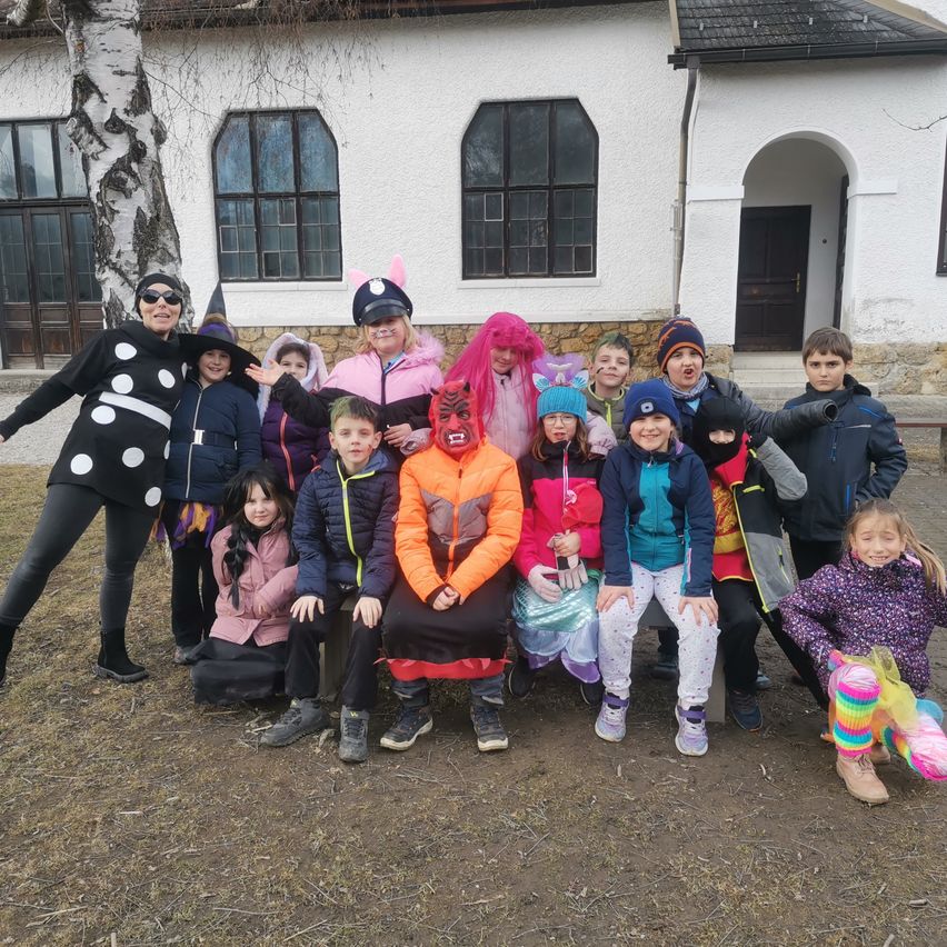 Eine Gruppe von Kindern und Erwachsenen in Kostümen posiert für ein Foto vor einem weißen Gebäude. Einige tragen Mützen, Brillen und Schminke im Gesicht.