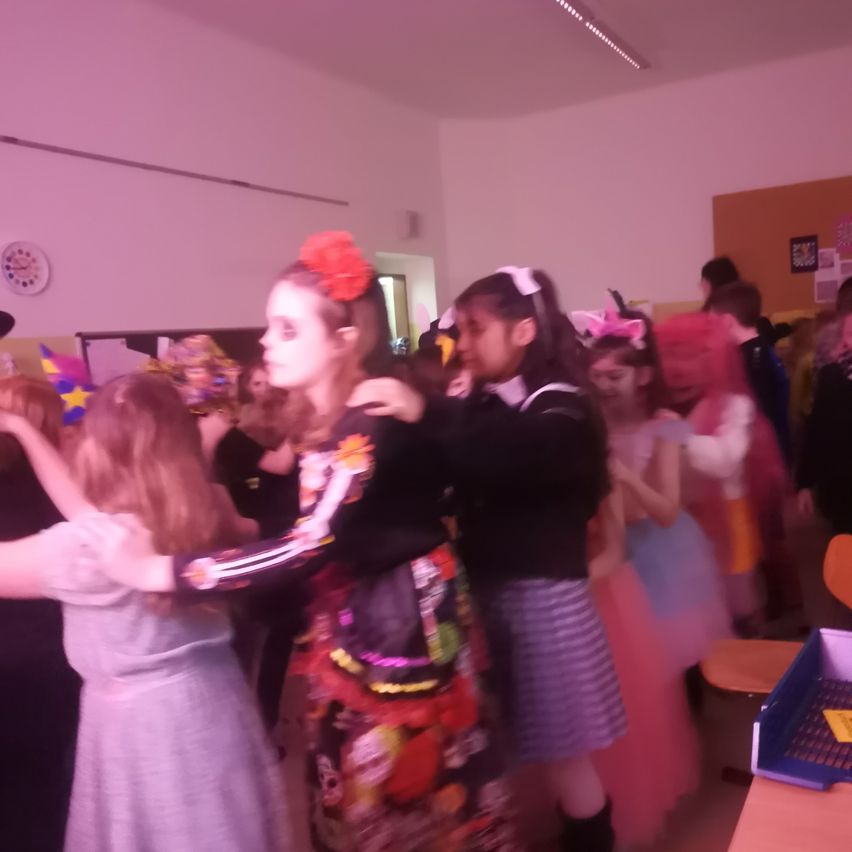 Mehrere Menschen in verschiedenen Kostümen tanzen gemeinsam in einem Raum. Eine Frau mit einer roten Blume im Haar steht in der Mitte.